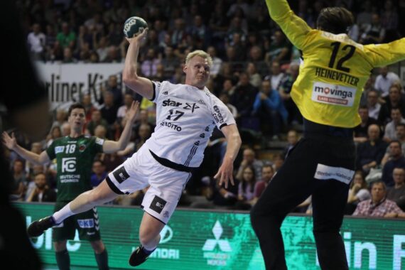 Ein Handballspieler in einem weißen Trikot springt, um den Ball zu schießen, während ein Torwart in Gelb die Arme zum Abwehren hebt. Die Aktion findet vor einer Menschenmenge in einer Halle statt.