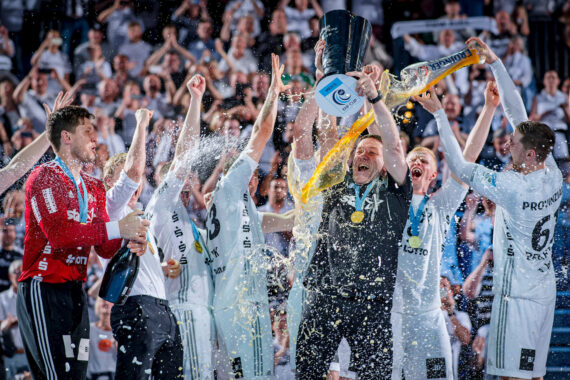 Eine jubelnde Handballmannschaft feiert den Sieg, hebt eine Trophäe in die Höhe und versprüht Champagner, während die Mannschaftskameraden jubeln, Konfetti fällt und die Fans im Hintergrund applaudieren.