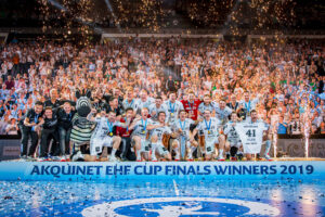 Ein jubelndes Handballteam in weißen Trikots feiert auf der Bühne mit einem Pokal in der Hand, umgeben von Konfetti, einem Zebramaskottchen und jubelnden Fans. Auf einem Transparent steht "AKQUINET EHF CUP FINALS WINNERS 2019".