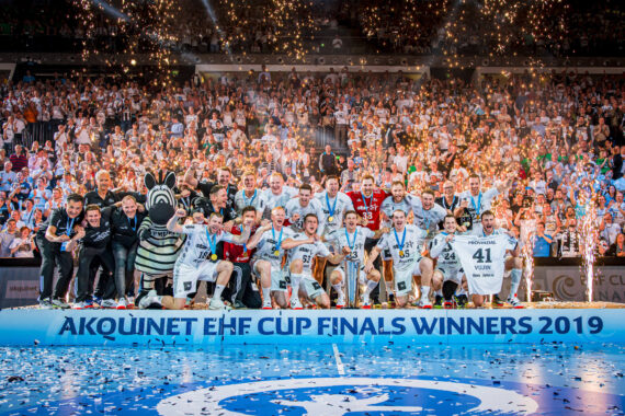 Ein jubelndes Handballteam in weißen Trikots feiert auf der Bühne mit einem Pokal in der Hand, umgeben von Konfetti, einem Zebramaskottchen und jubelnden Fans. Auf einem Transparent steht "AKQUINET EHF CUP FINALS WINNERS 2019".