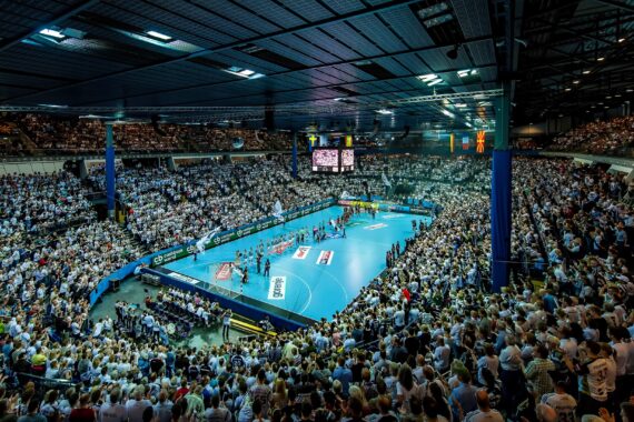 Eine überfüllte Halle mit Tausenden von Zuschauern, die ein Handballspiel auf einem hellblauen Spielfeld verfolgen, umgeben von Werbebannern und stimmungsvoller Beleuchtung.