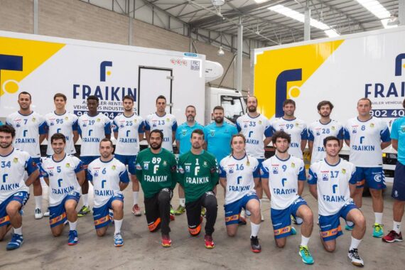 Eine Männerhandballmannschaft in weiß-blauen Trikots posiert für ein Gruppenfoto vor zwei gelb-weißen Fraikin-LKWs in einer Lagerhalle. Trainer und Mitarbeiter in Blau stehen an den Seiten.