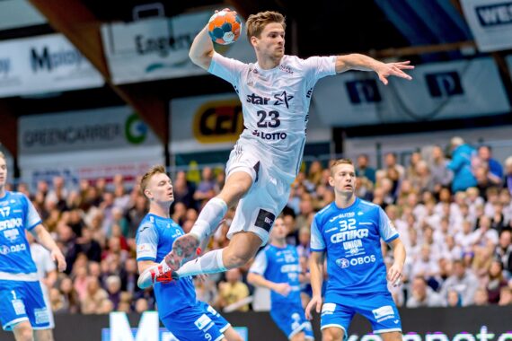 Ein Handballspieler in einem weißen Trikot springt in der Luft, um den Ball zu werfen, während seine blau gekleideten Gegner in einer überfüllten Halle zuschauen und versuchen, ihn zu verteidigen.