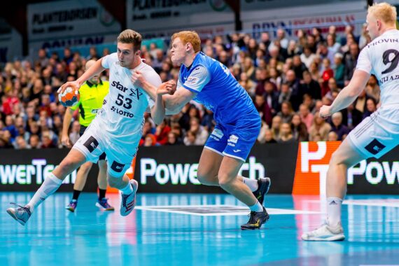 Zwei männliche Handballspieler in Aktion; der eine im weißen Trikot dribbelt den Ball, während ein Spieler in Blau versucht, ihn zu blocken. Eine Menschenmenge schaut von der Tribüne im Hintergrund zu.