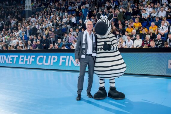 Ein Mann in Anzug und Schal steht lächelnd neben einer Person in einem Zebra-Maskottchenkostüm auf einem Sportplatz, mit einer großen Zuschauermenge im Hintergrund.