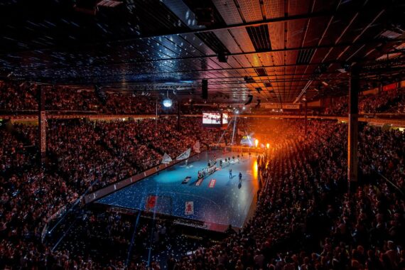 Eine große, mit Zuschauern gefüllte Halle umgibt ein hell erleuchtetes Handballfeld, Konfetti liegt auf dem Boden und Scheinwerfer beleuchten die Spieler während einer Veranstaltung oder Zeremonie.