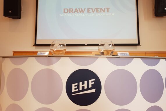 Ein Konferenztisch mit zwei Glasschalen mit der Aufschrift TEAMS und GROUPS steht vor einem Bildschirm mit der Aufschrift DRAW EVENT 2018/2019 MENS EHF CUP - GROUP PHASE. Auf dem Tisch befindet sich ein EHF-Logo.