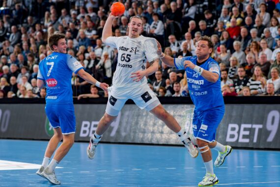 Ein Handballspieler in einem weißen Trikot springt, um zu schießen, während er den Ball in der Hand hält, während zwei Gegner in blauen Trikots versuchen, ihn auf einem Hallenplatz zu blockieren, während im Hintergrund eine Menschenmenge zusieht.