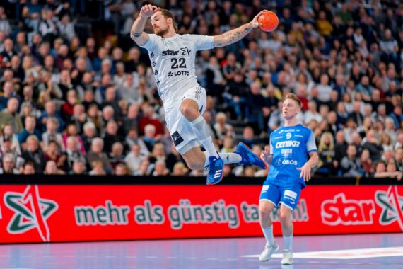 Ein Handballspieler in einem weißen Trikot springt, um den Ball zu werfen, während ein Spieler in Blau hinter ihm steht. Die Szene spielt sich auf einem Hallenplatz mit Zuschauern und einer roten Werbung im Hintergrund ab.