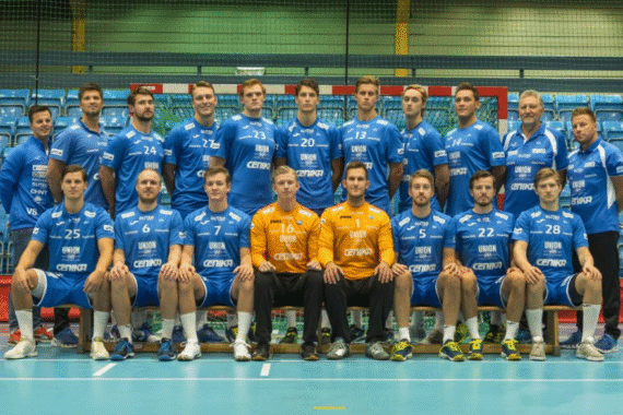 Ein Männersportteam in blauen Uniformen stellt sich in einer Halle für ein Gruppenfoto auf. Vier Personen in orangefarbenen Uniformen sitzen in der vorderen Mitte, der Rest steht und sitzt auf Bänken hinter ihnen in zwei Reihen.