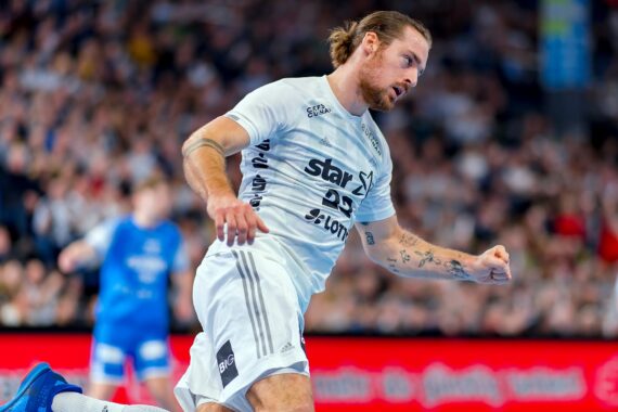 Ein Handballspieler in einem hellblauen Trikot jubelt auf dem Spielfeld und ballt die Faust, während im Hintergrund eine unscharfe Menschenmenge und andere Spieler in blauen Trikots zu sehen sind.