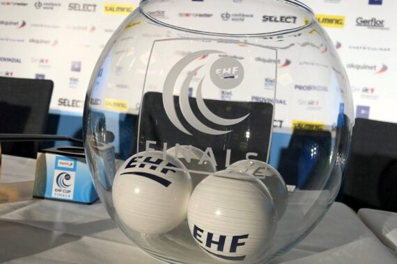 Eine klare Glasschale mit der Aufschrift EHF FINALS enthält zwei weiße Kugeln mit EHF-Logos. Die Schale steht auf einem Tisch, auf dem im Hintergrund Sponsorenlogos zu sehen sind.