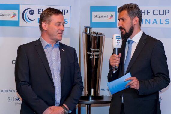 Zwei Männer in Anzügen stehen bei einem EHF-Pokalfinale neben einer großen Trophäe. Der eine hält ein Mikrofon und blaue Notizkarten in der Hand und scheint den anderen zu interviewen, während im Hintergrund Veranstaltungsbanner zu sehen sind.