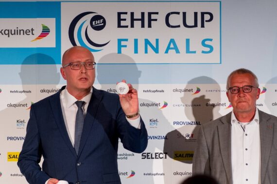 Zwei Männer in Anzügen stehen vor einem EHF CUP FINALS-Schild; einer hält einen kleinen Ball mit einem Schriftzug darauf hoch. Mehrere Sponsorenlogos zieren die Wand im Hintergrund.