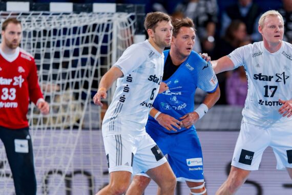 Zwei Handballspieler in weißen Trikots verteidigen gegen einen Spieler in einem blauen Trikot in Tornähe, während ein Torwart in einem roten Trikot vom Torraum aus zusieht. Alle Spieler scheinen auf das Spiel konzentriert zu sein.