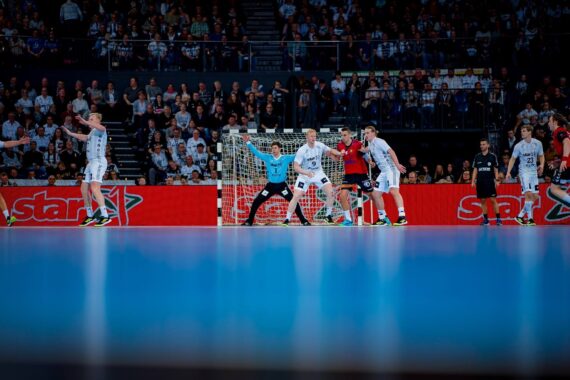 Handballspieler in weißen und roten Trikots treten vor einer überfüllten Arena an, der Torwart steht vor dem Tor bereit. Der Boden ist blau und im Hintergrund ist eine leuchtend rote Werbung zu sehen.