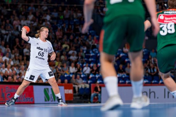 Ein Handballspieler in einem weißen Trikot bereitet sich darauf vor, den Ball zu werfen, während er zwei gegnerischen Spielern in grünen Trikots gegenübersteht.