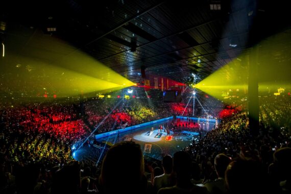 Eine überfüllte Sporthalle mit bunten Lichtern, die auf die Zuschauer und das Basketballfeld scheinen und eine dramatische und festliche Atmosphäre während einer Veranstaltung schaffen.
