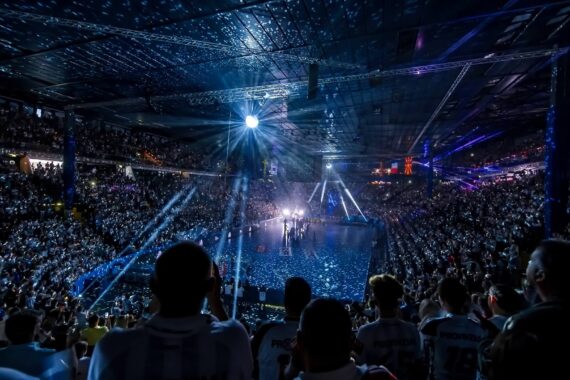 Ein großes Hallenstadion mit einer jubelnden Menge, beleuchtet von hellen Scheinwerfern und blauen Bühnenlichtern, die sich auf das Zentrum konzentrieren und eine festliche und energiegeladene Atmosphäre schaffen.