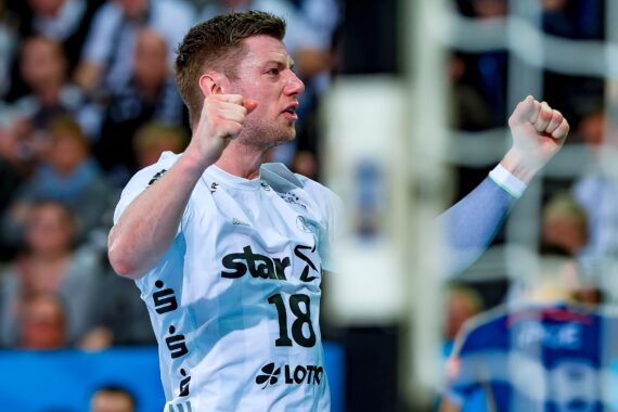 Ein Handballspieler in einem weißen Trikot mit der Nummer 18 jubelt mit geballten Fäusten, umgeben von einer unscharfen Menschenmenge im Hintergrund.