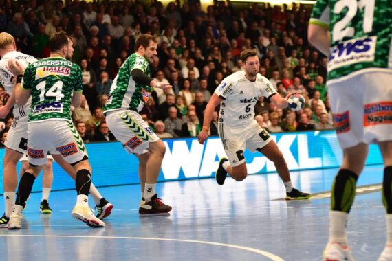Ein Handballspiel der Männer in Aktion: Ein Spieler in einem weißen Trikot dribbelt den Ball in Richtung Tor, umgeben von Verteidigern in grün-weißen Trikots. Die Zuschauer füllen die Tribünen im Hintergrund.