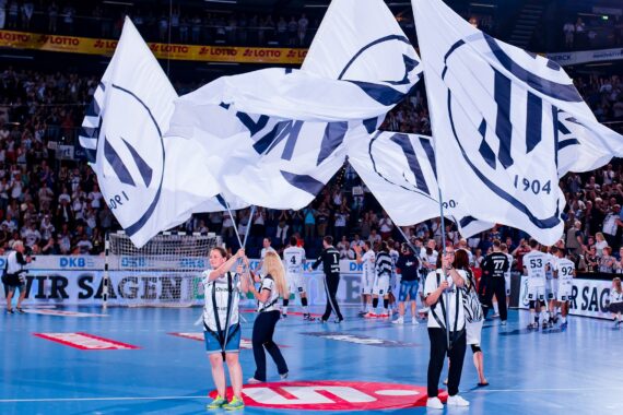 Menschen schwenken große weiße Fahnen mit einem schwarzen Logo auf einem Hallensportplatz mit einer jubelnden Menge auf der Tribüne. Im Hintergrund sind Spieler und Schiedsrichter zu sehen, was auf eine Sportveranstaltung hindeutet.