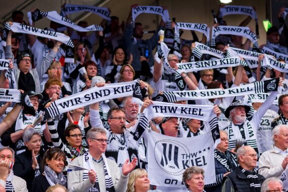 Eine große Menge begeisterter Fans in Schwarz und Weiß hält bei einer Sportveranstaltung Schals mit der Aufschrift WIR SIND KIEL und THW KIEL hoch, um ihre Mannschaft anzufeuern und zu unterstützen.