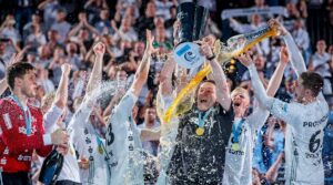 Eine jubelnde Handballmannschaft feiert auf dem Spielfeld, hebt einen Pokal in die Höhe und verspritzt Getränke, während im Hintergrund Konfetti und begeisterte Fans zu sehen sind. Die Spieler tragen weiße Trikots und Medaillen und jubeln mit erhobenen Armen.