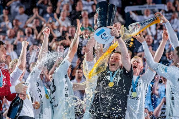 Eine jubelnde Handballmannschaft feiert auf dem Spielfeld, hebt einen Pokal in die Höhe und verspritzt Getränke, während im Hintergrund Konfetti und begeisterte Fans zu sehen sind. Die Spieler tragen weiße Trikots und Medaillen und jubeln mit erhobenen Armen.