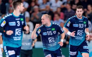 Drei männliche Handballspieler in blauen Trikots stehen auf dem Spielfeld, wobei einer in der Mitte eine feierliche Geste mit der Faust macht. Im Hintergrund ist eine unscharfe Zuschauermenge zu sehen.
