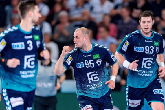 Drei männliche Handballspieler in blauen Trikots stehen auf dem Spielfeld, wobei einer in der Mitte eine feierliche Geste mit der Faust macht. Im Hintergrund ist eine unscharfe Zuschauermenge zu sehen.