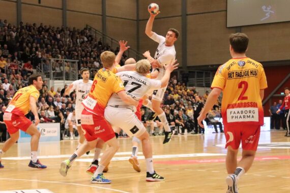 Eine Gruppe von männlichen Handballspielern ist auf einem Hallenplatz in Aktion. Ein Spieler in Weiß springt, um den Ball zu werfen, während mehrere Gegner in Gelb und Rot versuchen, ihn zu blockieren. Die Zuschauer füllen die Tribünen im Hintergrund.