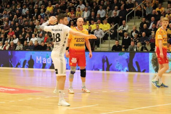 Ein Handballspieler in einem weißen Trikot bereitet sich auf den Wurf vor, während ein Gegner in einem orangefarbenen Trikot den Ball verteidigt. Das Spiel findet in einer Halle statt und wird von vielen Zuschauern auf der Tribüne verfolgt.