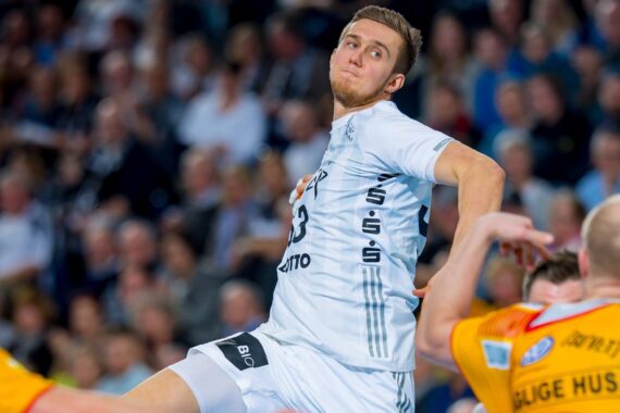Ein Handballspieler in einem weißen Trikot springt während eines Spiels in der Luft und bereitet sich darauf vor, den Ball zu werfen, während gegnerische Spieler in gelben Trikots versuchen, ihn zu blockieren. Im Hintergrund ist eine unscharfe Zuschauermenge zu sehen.