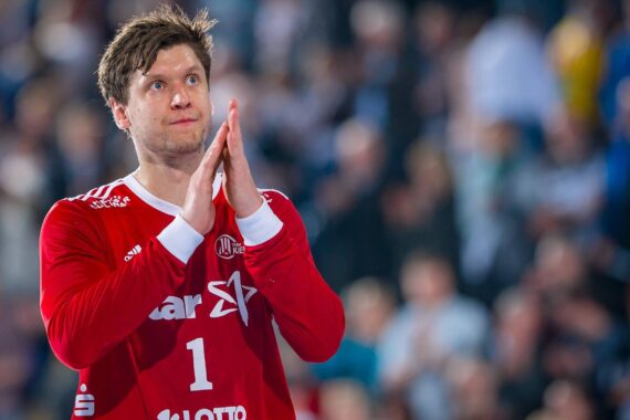 Ein männlicher Handballspieler in einem roten Trikot mit der Nummer 1 und Sponsorenlogos steht auf einem Spielfeld, faltet die Hände und blickt nach oben. Im Hintergrund ist eine unscharfe Zuschauermenge zu sehen.