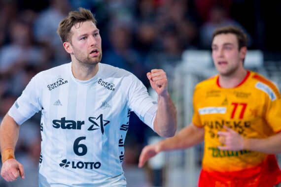 Ein männlicher Handballspieler in einem weißen Trikot mit der Nummer 6 ballt beim Jubeln die Faust, während ein anderer Spieler in einem gelben Trikot im Hintergrund ein Spiel verfolgt.