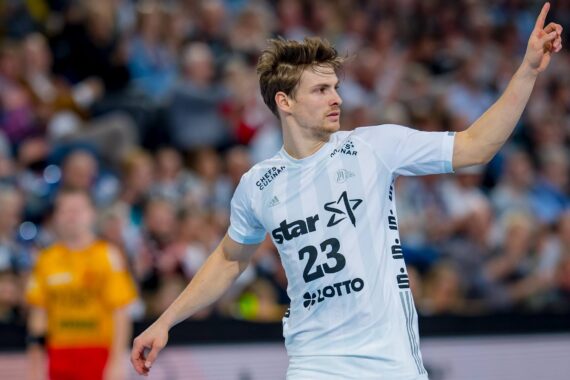 Ein männlicher Handballspieler, der ein weißes Trikot mit der Nummer 23 trägt, hebt während eines Spiels seinen rechten Arm und zeigt nach oben, während im Hintergrund eine unscharfe Menschenmenge zu sehen ist.