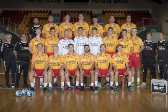 Eine Herrenhandballmannschaft und ihre Betreuer posieren für ein Gruppenfoto auf einem Hallenplatz. Die Spieler tragen gelbe und rote Trikots mit der Aufschrift Fynske Bank auf der Vorderseite, die Trainer und Mitarbeiter tragen schwarz oder weiß. Drei Handbälle liegen auf dem Boden vor ihnen.