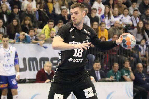 Ein männlicher Handballspieler in einem schwarzen Trikot mit der Nummer 18 bereitet sich darauf vor, den Ball während eines Spiels zu werfen, wobei im Hintergrund Zuschauer und andere Spieler zu sehen sind.