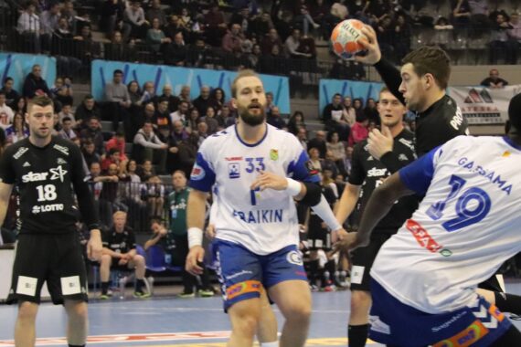 Ein Handballspieler in Schwarz springt, um zu schießen, während zwei Verteidiger in Weiß versuchen, ihn in einem Hallenstadion mit einer großen Zuschauermenge zu blockieren. Andere Spieler und Schiedsrichter sind im Hintergrund zu sehen.
