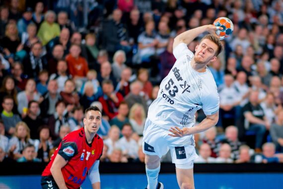 Ein männlicher Handballspieler in einem weißen Trikot springt, um den Ball zu werfen, während ein Spieler in einem roten Trikot aufmerksam zusieht; im Hintergrund ist eine Zuschauermenge zu sehen.