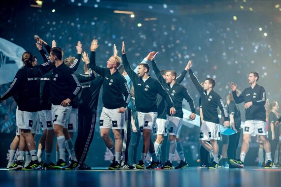 Eine Sportmannschaft in schwarz-weißen Trikots klatscht sich ab, als sie in einer Arena mit hellen Lichtern und einer jubelnden Menge im Hintergrund vorgestellt wird.
