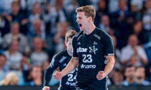Ein männlicher Handballspieler in einem schwarzen Trikot mit der Nummer 23 jubelt leidenschaftlich auf dem Spielfeld, ballt die Fäuste und schreit, während im Hintergrund eine verschwommene Menschenmenge zu sehen ist.