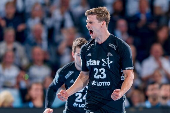 Ein männlicher Handballspieler in einem schwarzen Trikot mit der Nummer 23 jubelt leidenschaftlich auf dem Spielfeld, ballt die Fäuste und schreit, während im Hintergrund eine verschwommene Menschenmenge zu sehen ist.