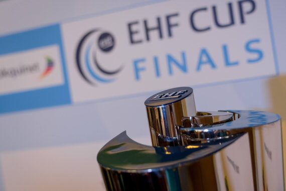 Eine Nahaufnahme einer silbernen Trophäe vor einem Schild mit der Aufschrift EHF CUP FINALS und einem blau-weißen Farbschema im Hintergrund.