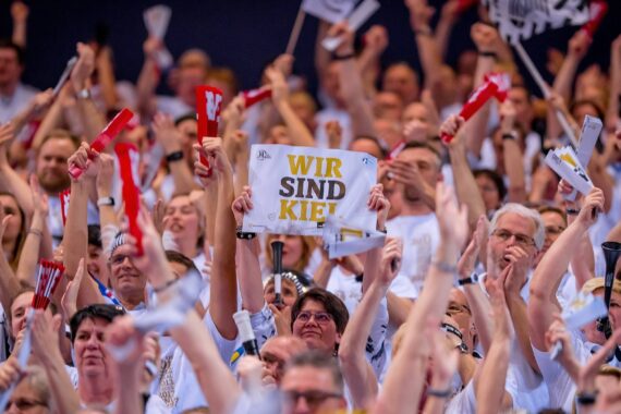 Eine große Menge begeisterter Fans in weißen Hemden jubelt und klatscht. Eine Person hält ein Schild hoch, auf dem in fetten gelben Buchstaben WIR SIND KIEL! steht.