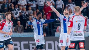 Zwei Handballspieler in blau-weißen Trikots feiern auf dem Spielfeld mit erhobenen Armen, während Spieler in rot-weißen Trikots und eine Menge Zuschauer im Hintergrund zusehen.