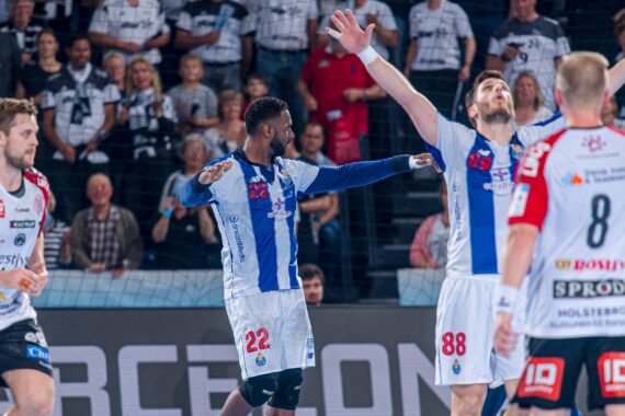Zwei Handballspieler in blau-weißen Trikots feiern auf dem Spielfeld mit erhobenen Armen, während Spieler in rot-weißen Trikots und eine Menge Zuschauer im Hintergrund zusehen.
