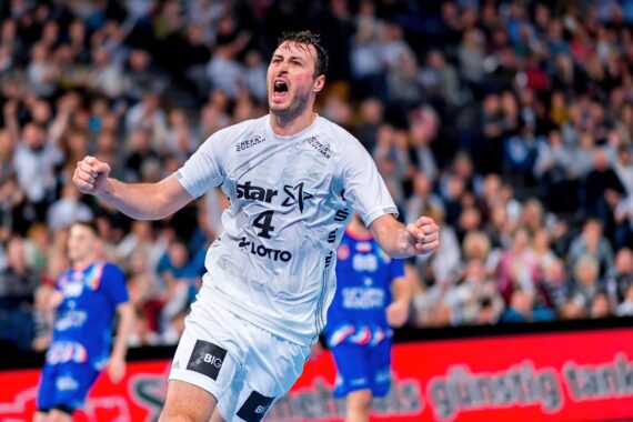 Ein Handballspieler, der ein weißes Trikot mit der Nummer 4 trägt, jubelt mit geballten Fäusten während eines Spiels, während im Hintergrund eine unscharfe Menge und andere Spieler zu sehen sind.
