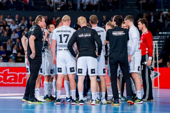 Eine Herren-Handballmannschaft in weiß-schwarzen Trikots sitzt während eines Spiels mit den Trainern auf einem Hallenplatz zusammen, während im Hintergrund ein Publikum zuschaut.